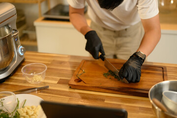 Close up of man wearing black gloves chopping herbs on a wooden cutting board