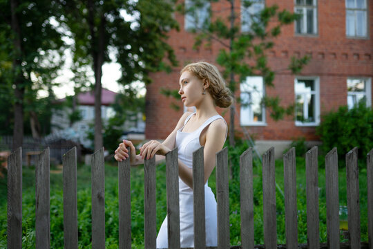 Girl in a white dress standing by a fence outdoors near a brick building