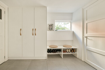 A bright and airy modern interior showcasing minimalist decor. The space features white cabinetry, a cozy shoe rack, and natural light streaming in through the window.