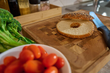 Sliced bagel on wooden cutting board with cherry tomatoes, broccoli and kitchen tools in...