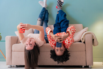 Caucasian and african american girls lying upside down on couch, smiling while using smartphones. A...