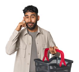 Hispanic male holding shopping basket in studio pointing temple with finger, thinking, focused on a task.