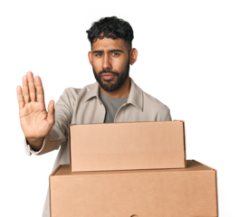 Hispanic male holding cardboard boxes in studio standing with outstretched hand showing stop sign, preventing you.