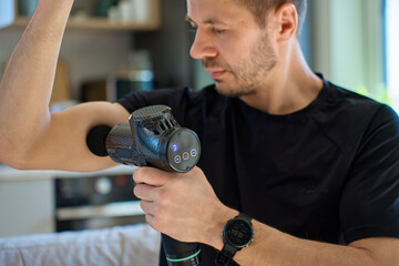Athletic man using percussion massage device indoors. Man applies massage gun to arm muscle while sitting in living room, recovery therapy after exercise. Concept of home fitness and relaxation © Lazy_Bear