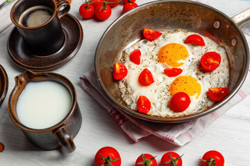 Delicious breakfast with fried eggs and cherry tomatoes in a pan, coffee and milk