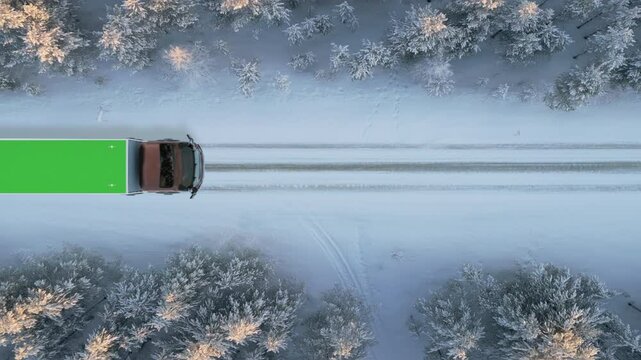 aerial top down view of cargo truck with green screen roof driving on the road covered of snow in the forest,shipping delivery concept,copyspace for advertising