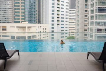 Young asian woman relaxing by pool at Kuala Lumpur hotel with view of surrounding skyscrapers, enjoying leisure time in vibrant urban setting.