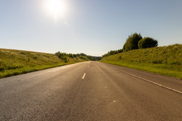 a highway in a rural area with a blue sky and green grass on the side of the road, The sun is shining in the sky