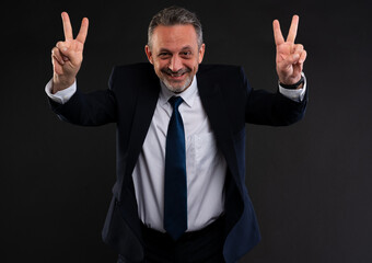 Smiling mature man in a suit making two peace or victory signs with both hands, expressing excitement, positivity and confidence in a studio setting.