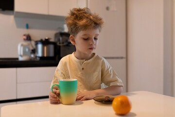 Young boy enjoys morning snack at home while sitting at kitchen table with colorful cup and fruit...