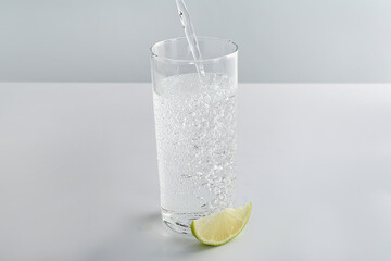 Pouring refreshing soda water into glass at white table against light background