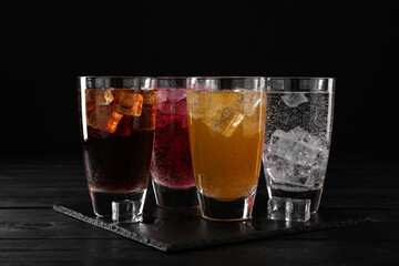 Refreshing soda water of different flavors with ice cubes in glasses on black wooden table