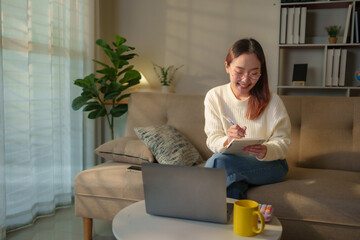 Asian freelancer writing on notepad while sitting on comfortable sofa near laptop and coffee, working remotely from home