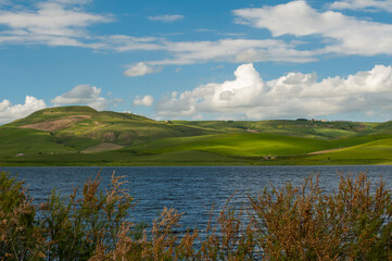 Alta Murgia, panorama del parco con prato , cielo azzurro e nuvole, lago e casolari. Il lago fra le...