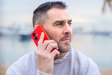 Caucasian man stands outdoors near water, talking on a smartphone with a serious look. His casual...
