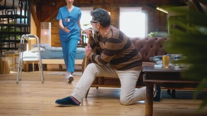 Female caregiver helps elderly 60s man to get up from the floor after a fall accident and mobility problems in the living room at home, he feels pains in his back and legs.