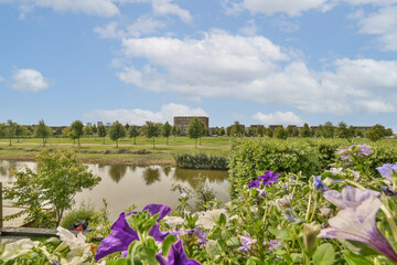 A picturesque view featuring vibrant flowers in the foreground, a tranquil pond, lush greenery, and a cloudy sky, creating a peaceful natural scene.