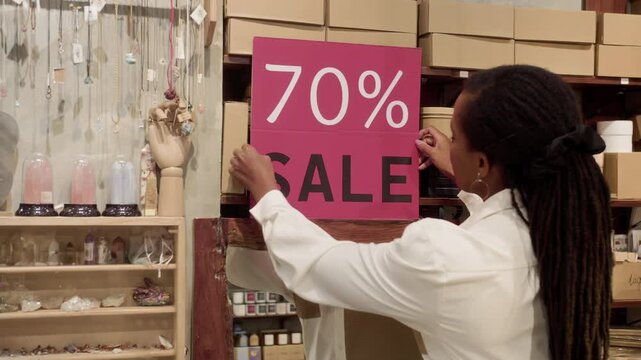 Medium static shot of young black woman organizing accessory store space placing sign with sale announcement