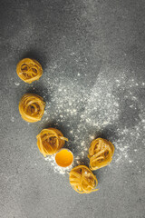 Top view of raw tagliatelle pasta nests on a floured surface and wooden board, with a cracked egg...