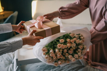 A wrapped gift with pink ribbon being handed over near a bouquet of roses and a cup of coffee on a...