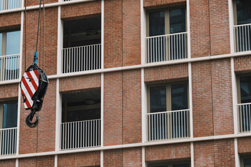 Industrial crane hook suspended in front of modern red brick facade, capturing tension between construction process and architectural completion.