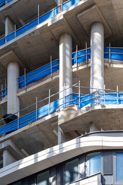 Detailed view of modern building facade under construction, with blue safety netting, exposed concrete columns and architectural grid elements.