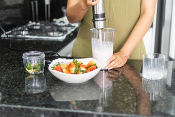 Woman preparing strawberry smoothie with hand blender in kitchen