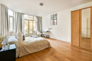 A bright bedroom featuring large windows, minimalistic decor, and wooden flooring, creating a serene atmosphere for relaxation and comfort.