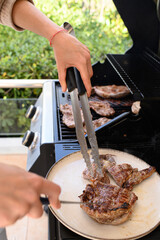Woman removing grilled meat from outdoor gas barbecue and placing it on a plate. Juicy, cooked steaks with grill marks. Summer BBQ, rustic food, home cooking in natural light.