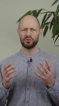 A guy blogger gives a speech and sometimes stutters, looking at the camera and gesturing, he is wearing a blue shirt and is in a studio next to a plant