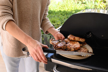 Woman removing grilled meat from outdoor gas barbecue and placing it on a plate. Juicy, cooked steaks with grill marks. Summer BBQ, rustic food, home cooking in natural light.