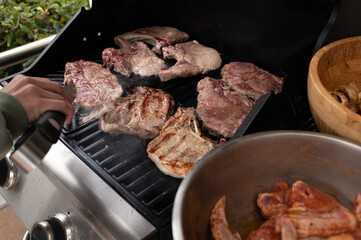 Hand holding tongs flipping grilled meat steaks on outdoor gas barbecue. Juicy, seared beef with grill marks. Summer cooking, backyard BBQ scene, high-protein lifestyle.