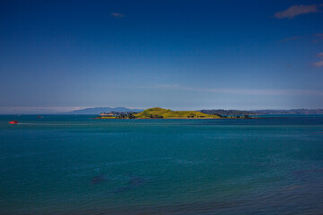 A stunning ocean view featuring a red ferry boat sailing near lush green islands in Auckland, New Zealand. The deep blue water contrasts beautifully with the vibrant landscape under a clear blue sky.