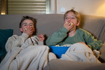 Two young brothers watching tv and eating popcorn on sofa at night