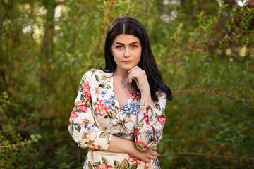 Fototapeta premium A confident woman wearing a stylish floral dress stands outdoors, surrounded by lush green foliage on a sunny day. The setting highlights natural beauty and the charm of fashionable attire.