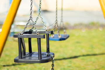 Empty children's hanging swing with a red plastic seat and silver metal chains, suspended from a sturdy metal frame. Outdoor setting with green grass and soft background blur.

