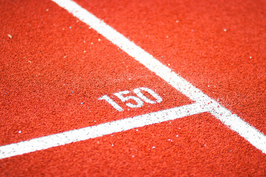 Distance lane markings on a red running track at an athletics stadium. White numbers and lines on textured rubber surface. Outdoor shot in daylight, focused on track detail.

