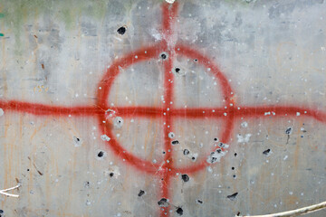 Damaged concrete wall with bullet holes and a red target painted over them. Old gray concrete surface with numerous bullet holes of various sizes and uneven edges. A red target in the form of a circle