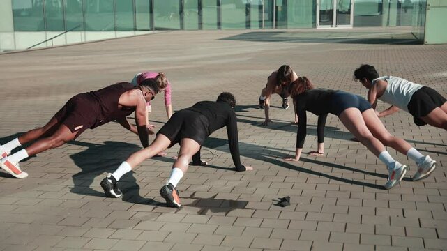 A multiethnic group of people wearing sportswear and sunglasses exercises on a paved floor in front of a glass building. They train their arms while standing in a push-up position in a circle.