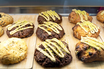 Close-up of assorted chocolate and vanilla cookies topped with pistachio crumbs and frosting, displayed on a wooden tray inside a bakery showcase.