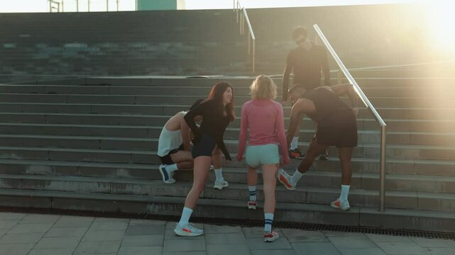 A group of five multiethnic fit people in their late 20s wearing athletic clothing warms up by doing stretches, on a wide concrete staircase, with a metal railing, outdoors in a city, on a sunny day.
