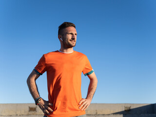 Sportsman resting after training wearing orange t-shirt and earphones under blue sky