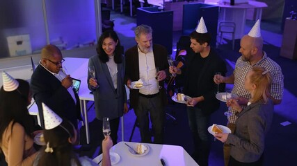 An Asian woman makes a toast in honor of a senior White man next to her, in a circle of her multiethnic colleagues, some in party hats, and then they clink glasses, in an office, at a retirement party