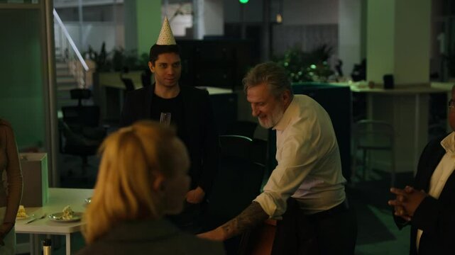 A smiling White man in his 60s thanks his multiethnic colleagues by bowing down and making a praying gesture, at his retirement party in an office with colorful changing lights, in the evening.