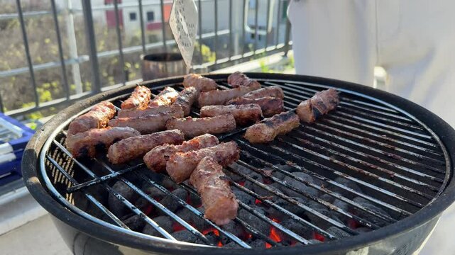 Cevapcici, small skinless sausages made of a mixture of ground meat, are grilling on a barbecue with glowing embers, and a spatula turns them over