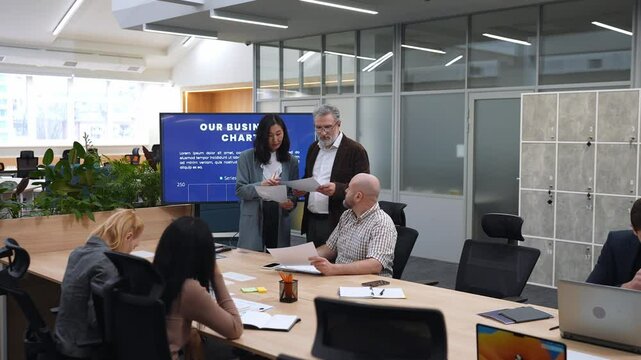 A middle-aged White businessman in his late 50s, talking to his mid-30s Asian female colleague about some papers as they stand near the table where their colleagues sit, in an office meeting room,
