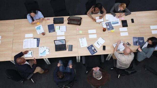 Several diverse colleagues work in an office with laptops, documents, and office supplies scattered on the table. Some take notes while others work on laptops or review materials in a meeting.
