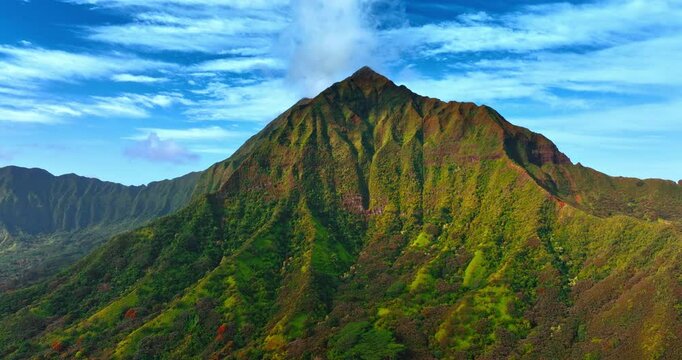 Flying closer to the peak of the stunning mountain in Koolau Range in the Oahu Island. Breath-taking rock covered with grass and bushes at the backdrop of blue sky.