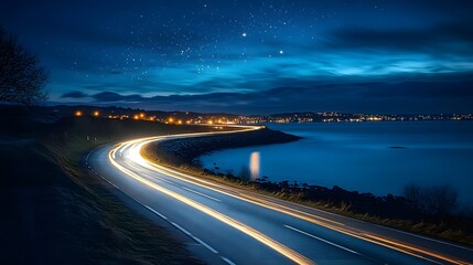 Obraz premium Night view of coastal highway with starburst streetlights beside calm ocean, twilight sky and distant town lights reflecting on water. 