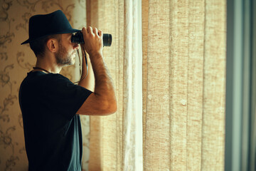 Man in fedora hat looking through binoculars spying from window